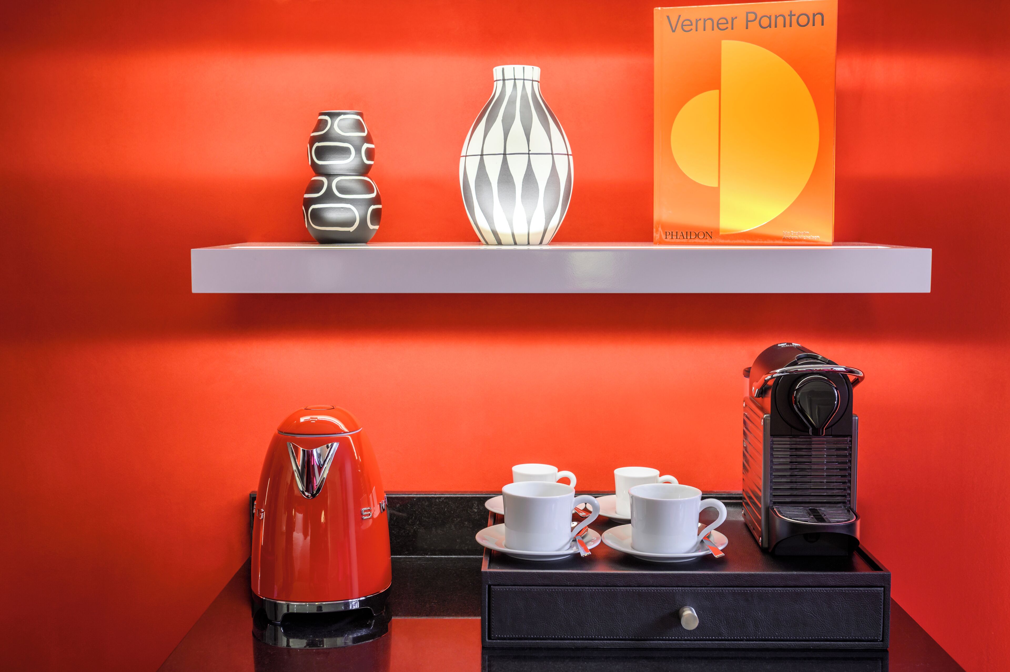 A hotel room with a red wall, a white shelf with decorative items, a red kettle, a coffee machine, and a black suitcase.