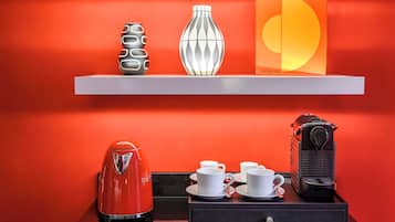 A hotel room with a red wall, a white shelf with decorative items, a red kettle, a coffee machine, and a black suitcase.