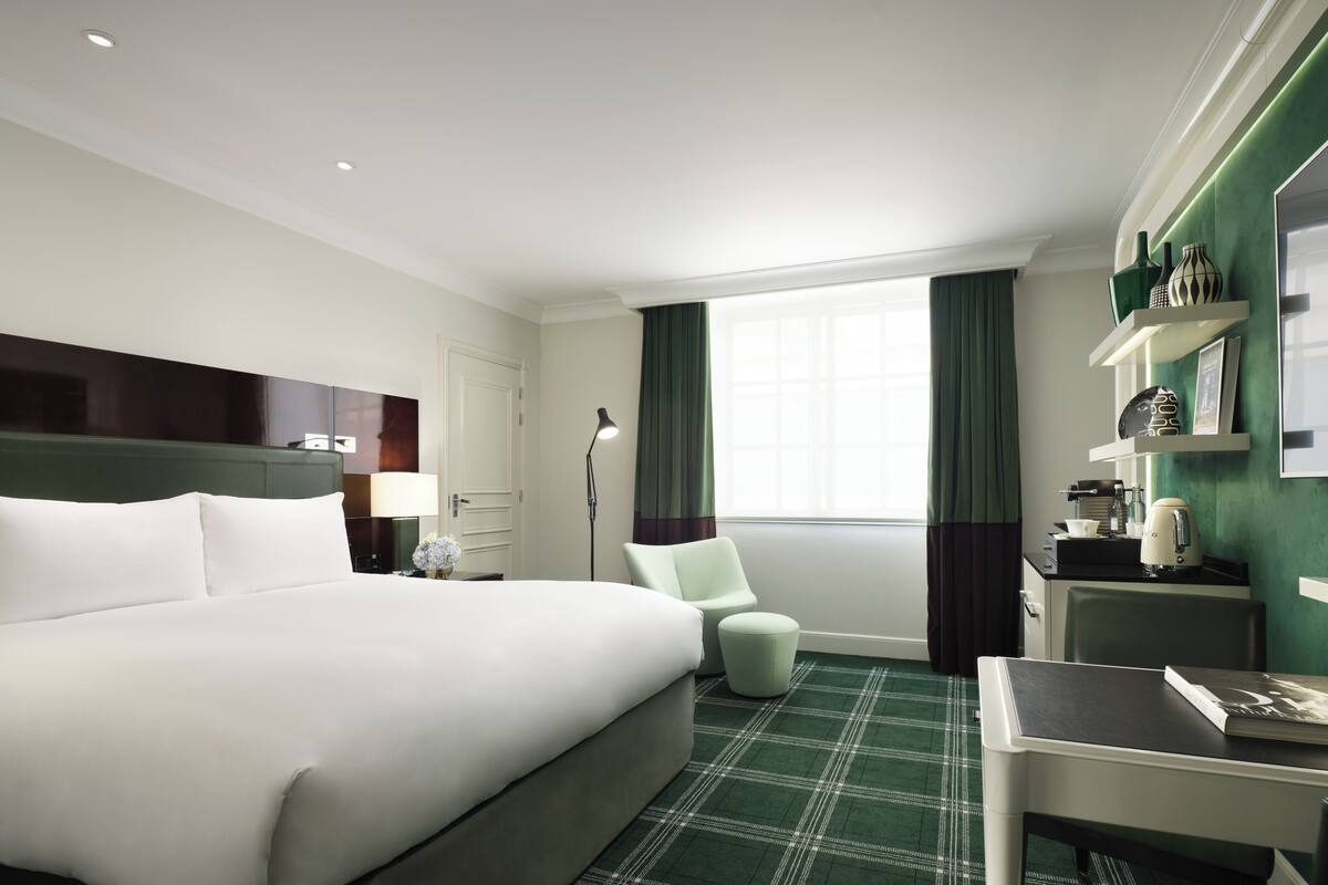 A hotel room with a large bed, a desk, a chair, a lamp, a coffee maker, and a window with curtains.