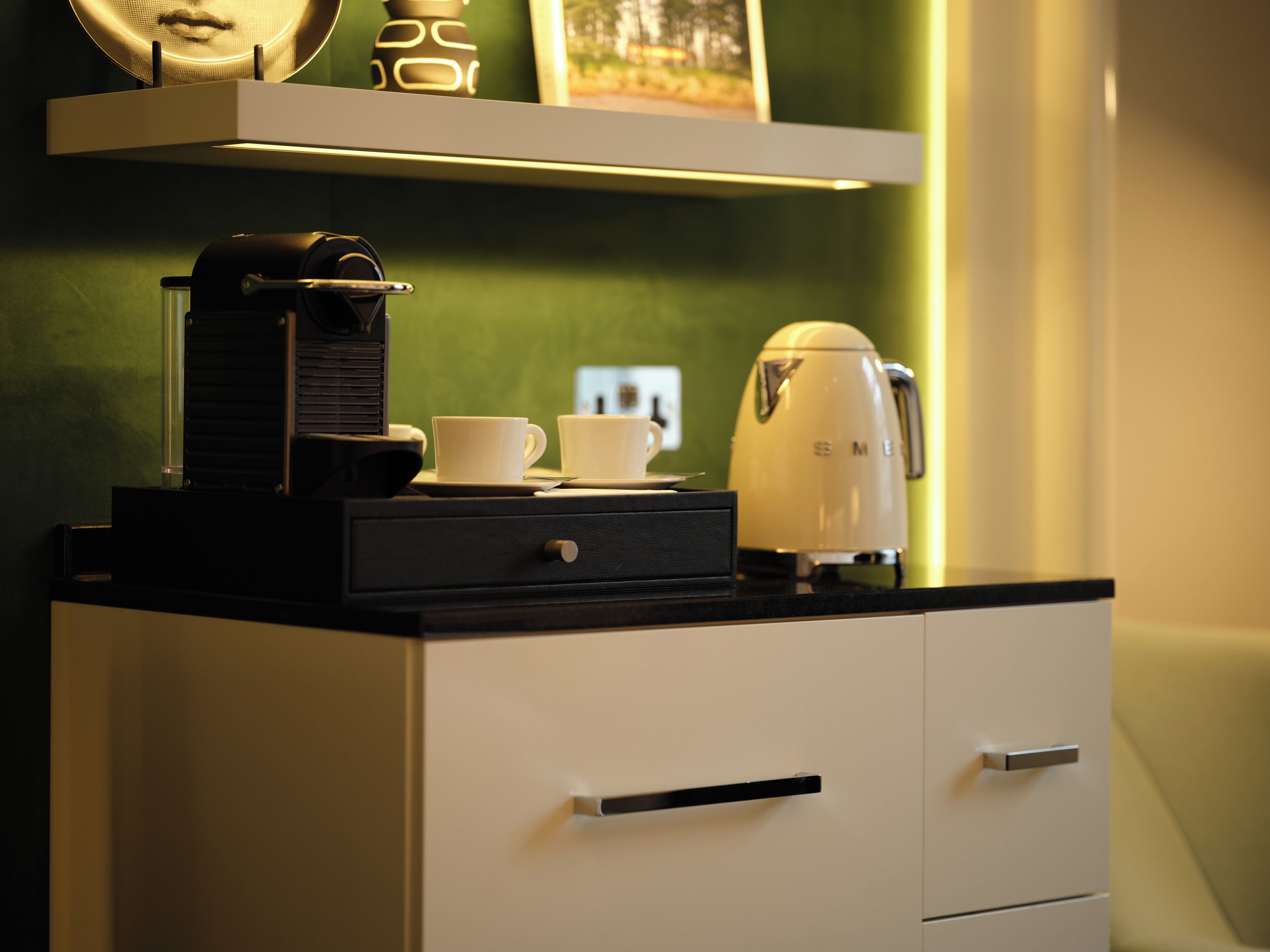 A kitchenette with a coffee maker, two cups, and a kettle on a countertop.