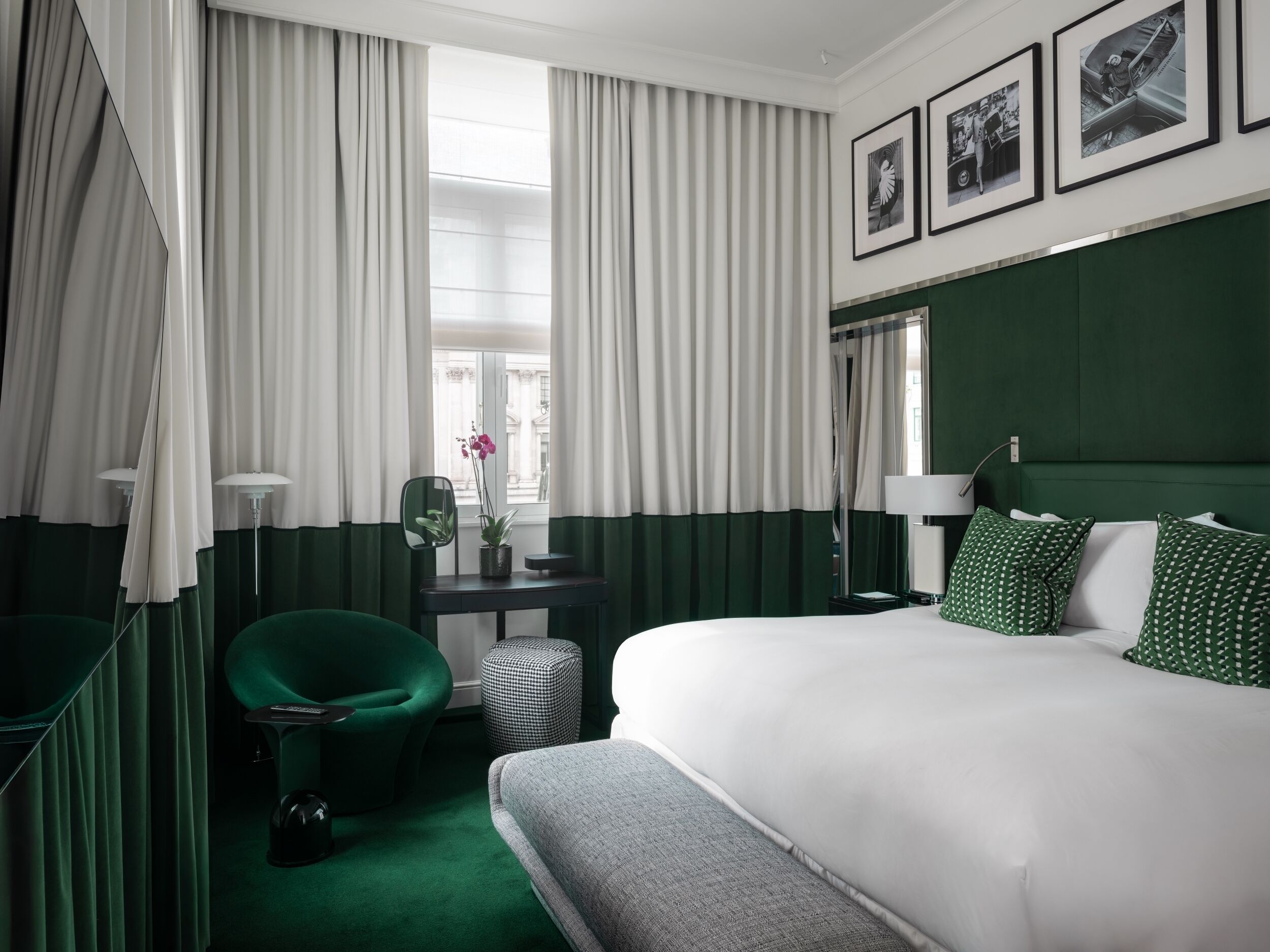 Una habitación de hotel con una cama grande, un sillón tapizado en verde, un escritorio pequeño con espejo y una ventana con cortinas de gasa.