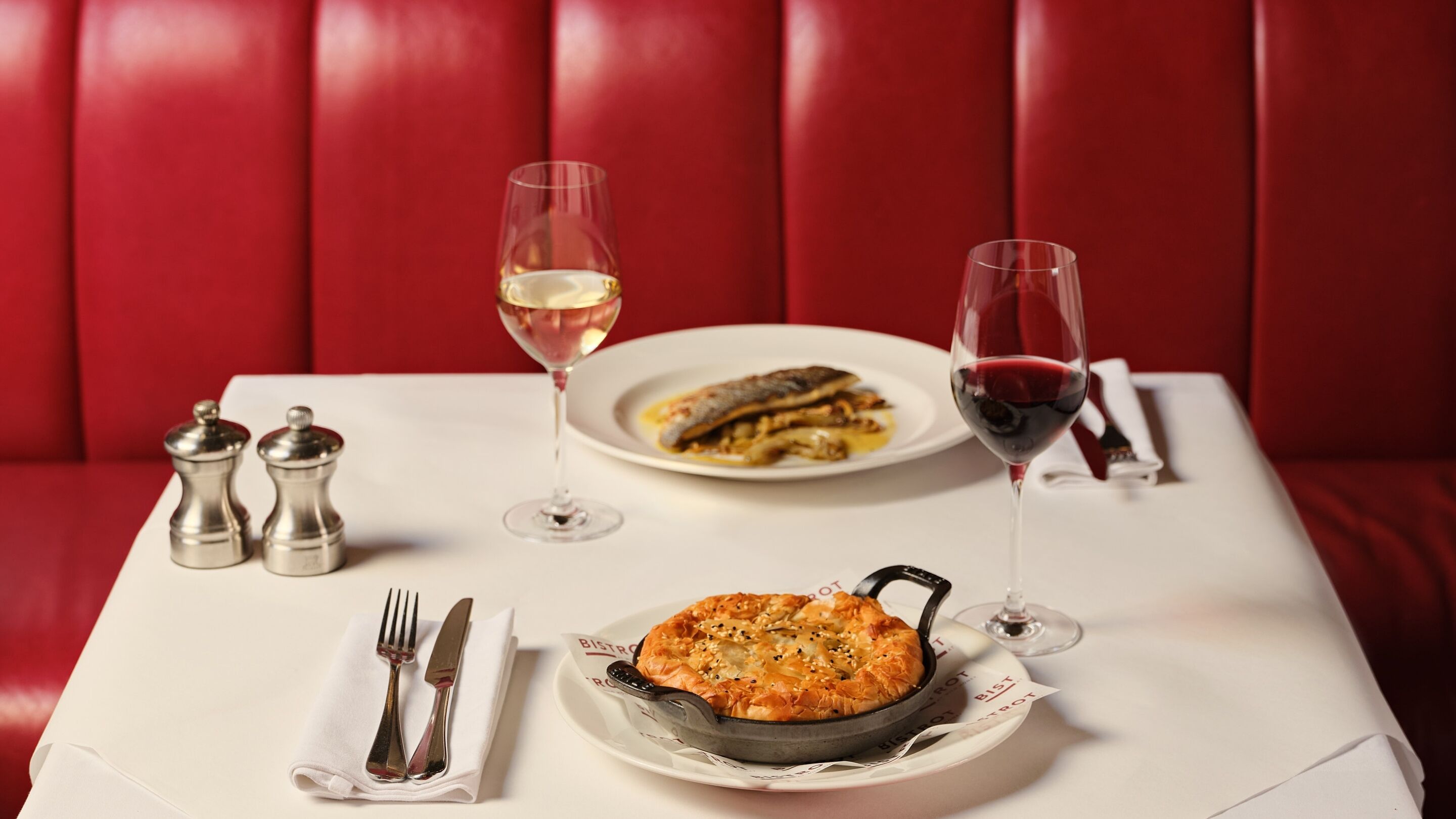 A dining table with a white tablecloth, two salt and pepper shakers, a plate of food, a cast iron skillet, a fork and knife set, and two wine glasses with white and red wine.