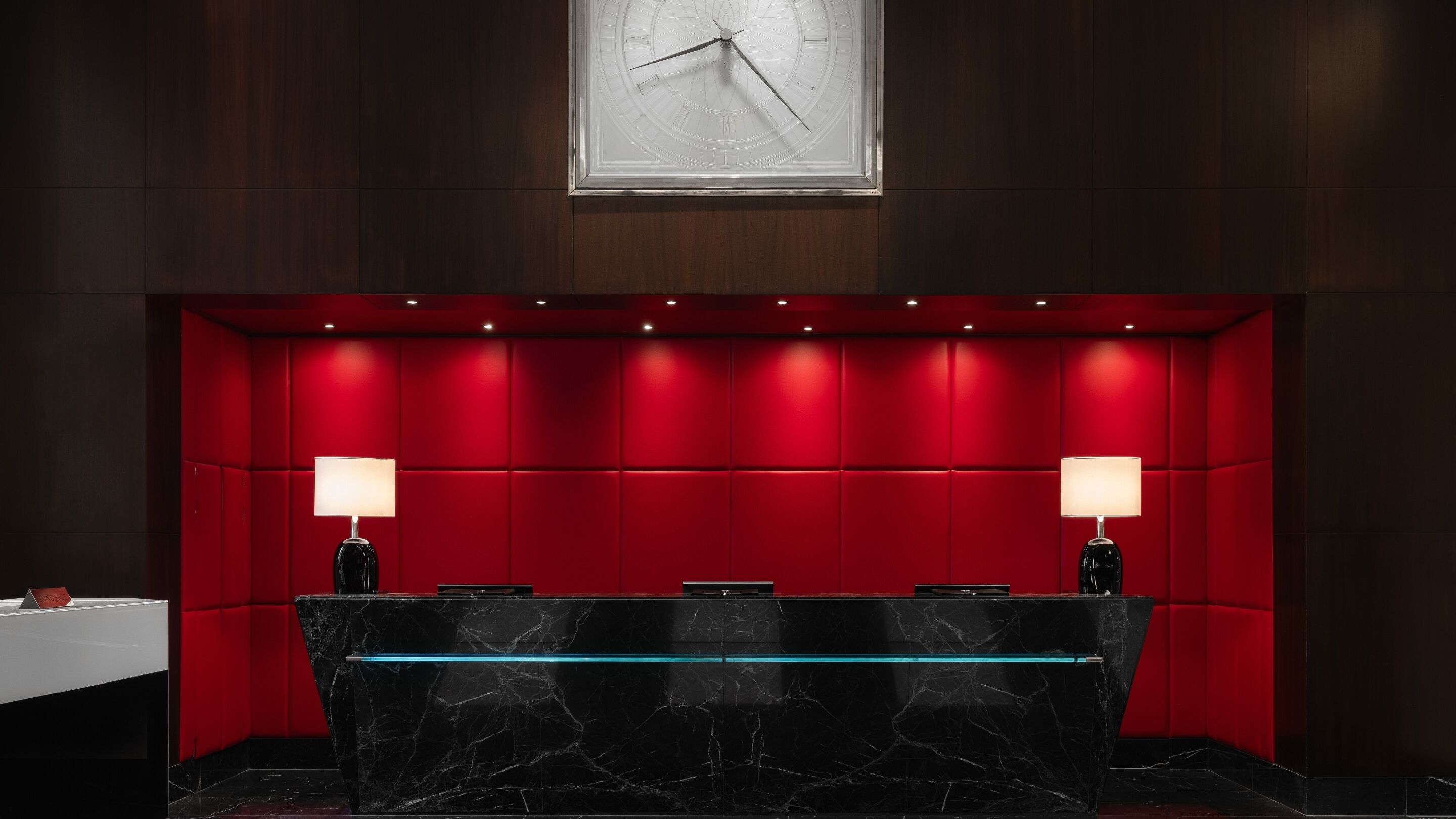 A reception desk with a marble top, red padded wall, two lamps, and a clock above.