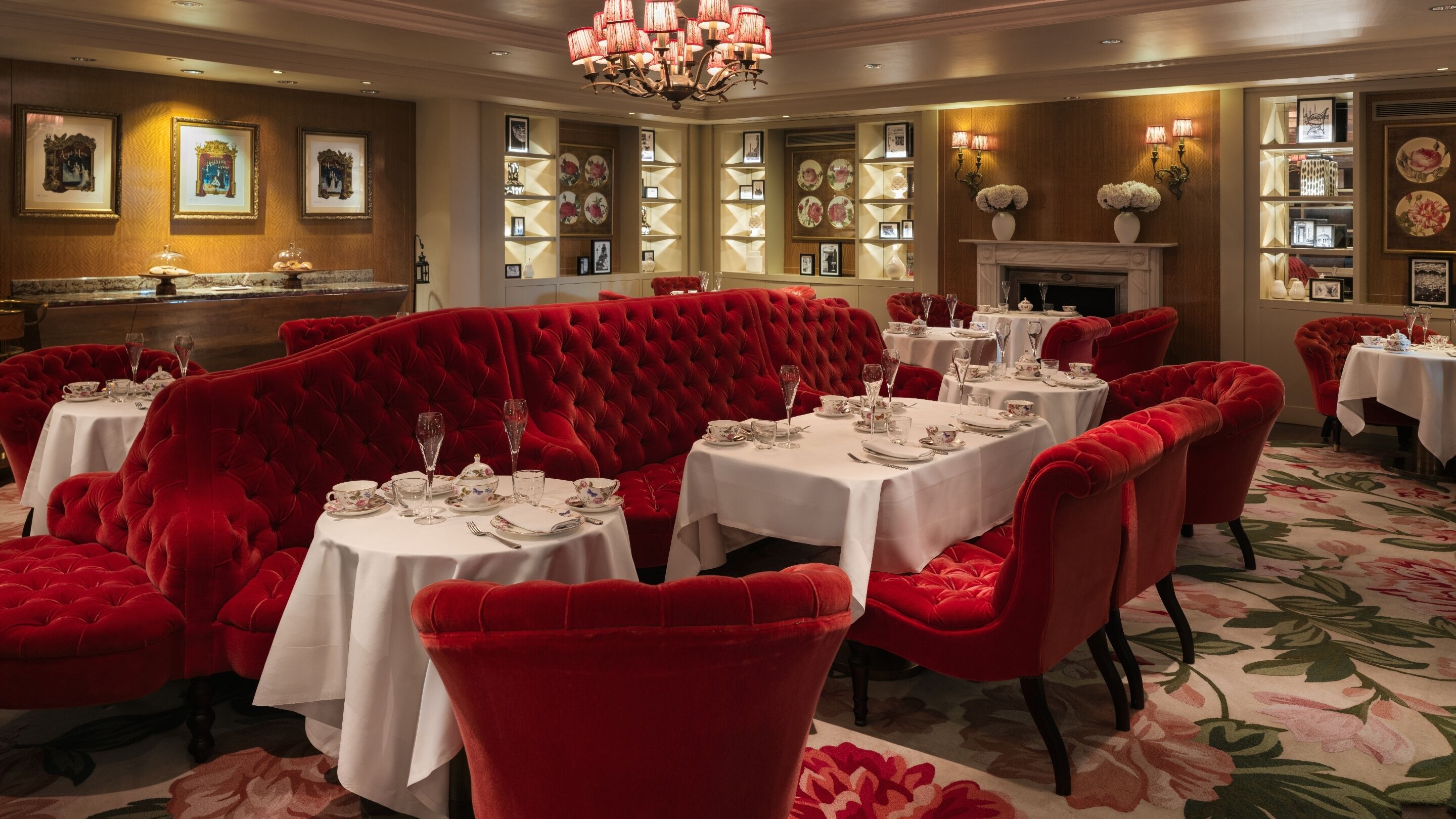 A dining area with red upholstered seating and white tablecloths, featuring a fireplace and framed artwork on the walls.