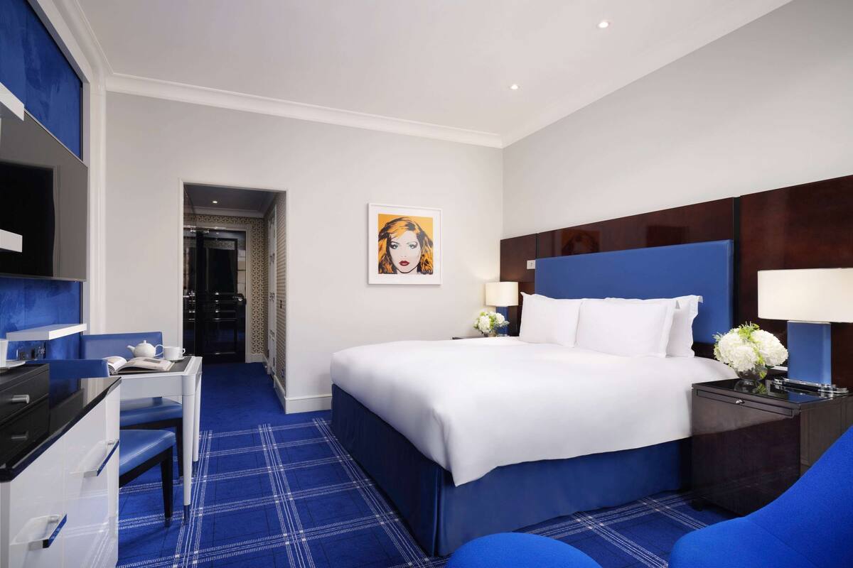 A hotel room with a bed, a desk, a chair, a TV, and a painting on the wall.