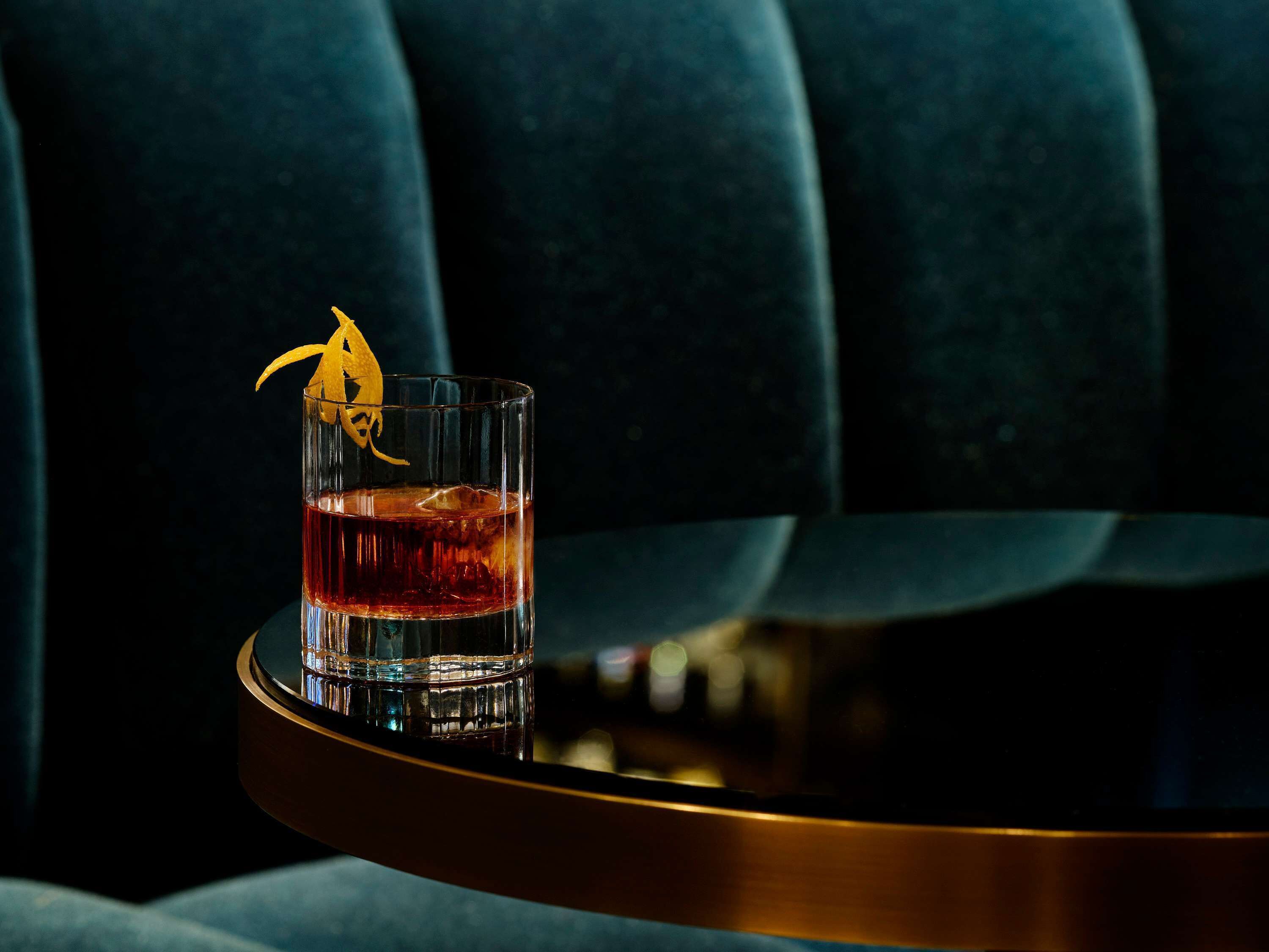 Un vaso de bebida color ámbar con un adorno amarillo sobre una mesa redonda frente a un fondo tapizado azul oscuro.