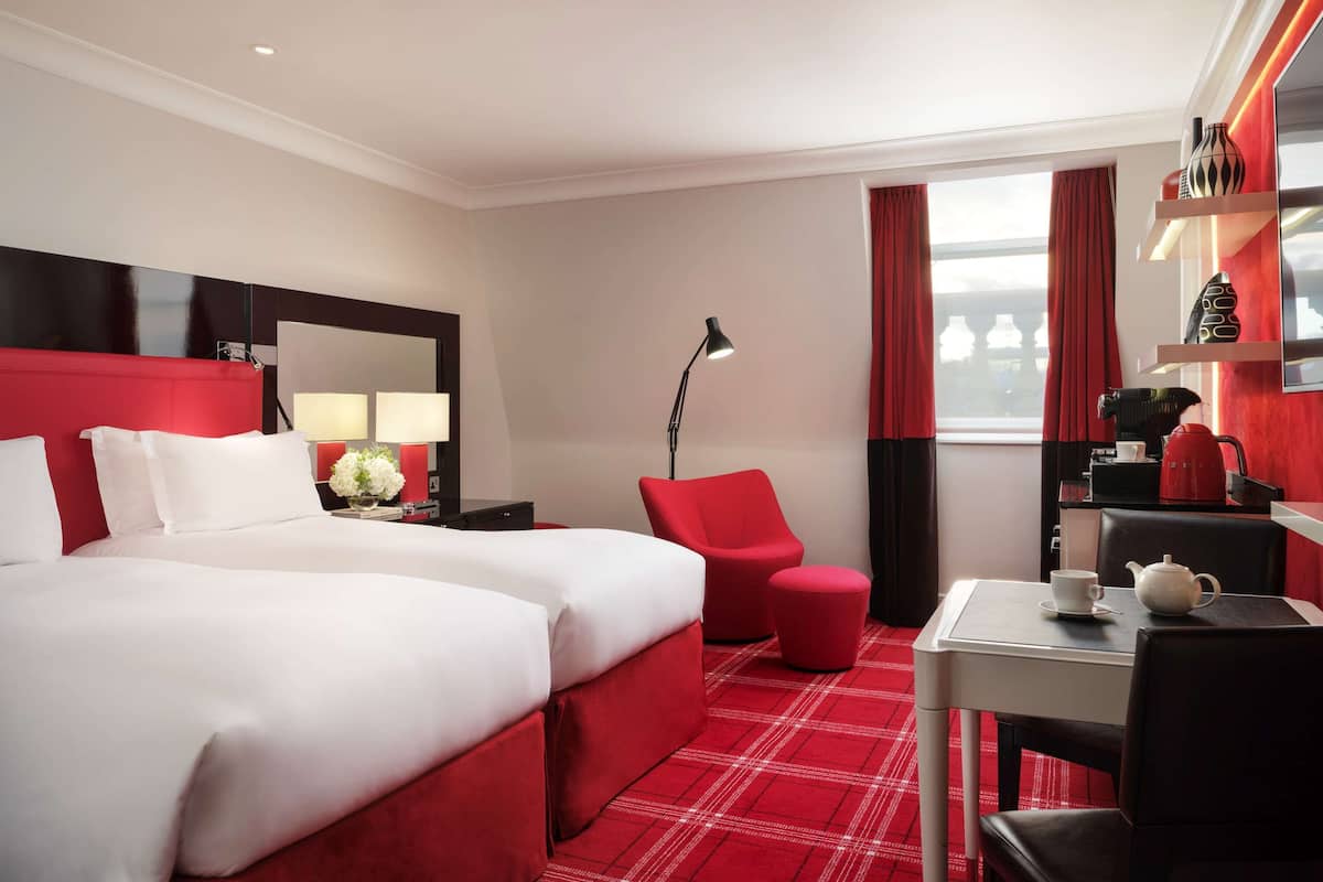 A hotel room with a large bed, a desk with a kettle, and a red chair.