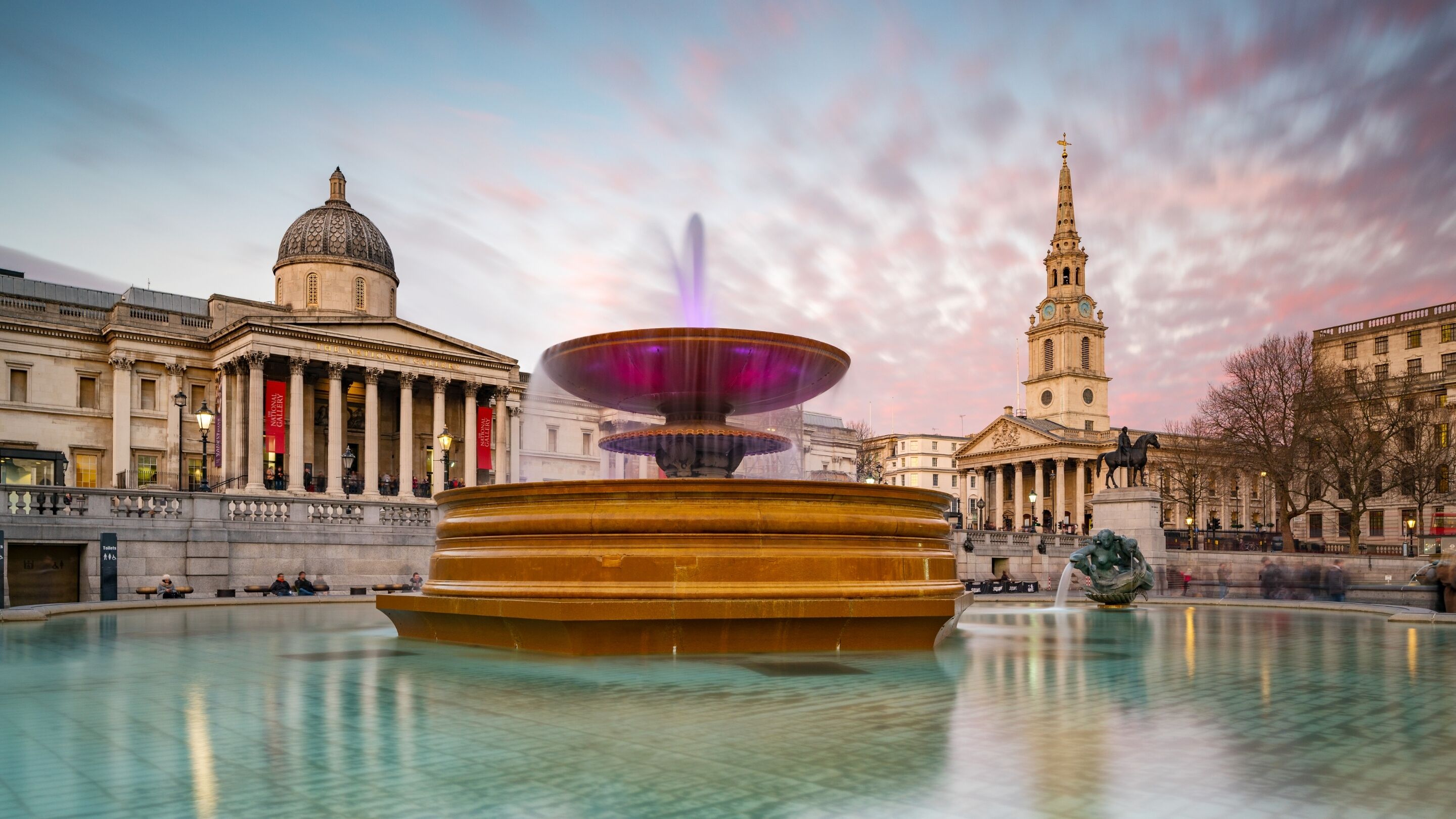 A historic plaza with a central fountain, classical architecture, and a prominent clock tower.