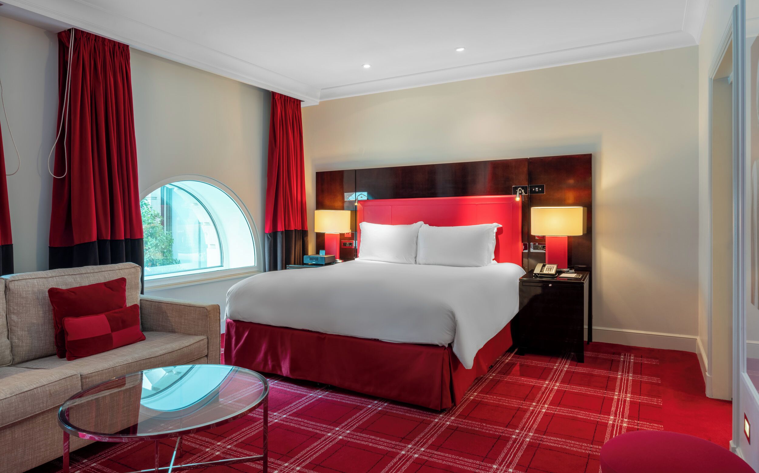 A hotel room with a large bed, two bedside tables with lamps, a sofa with a red cushion, a glass coffee table, and a window with red curtains.