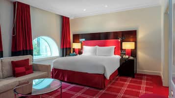 A hotel room with a large bed, two bedside tables with lamps, a sofa with a red cushion, a glass coffee table, and a window with red curtains.
