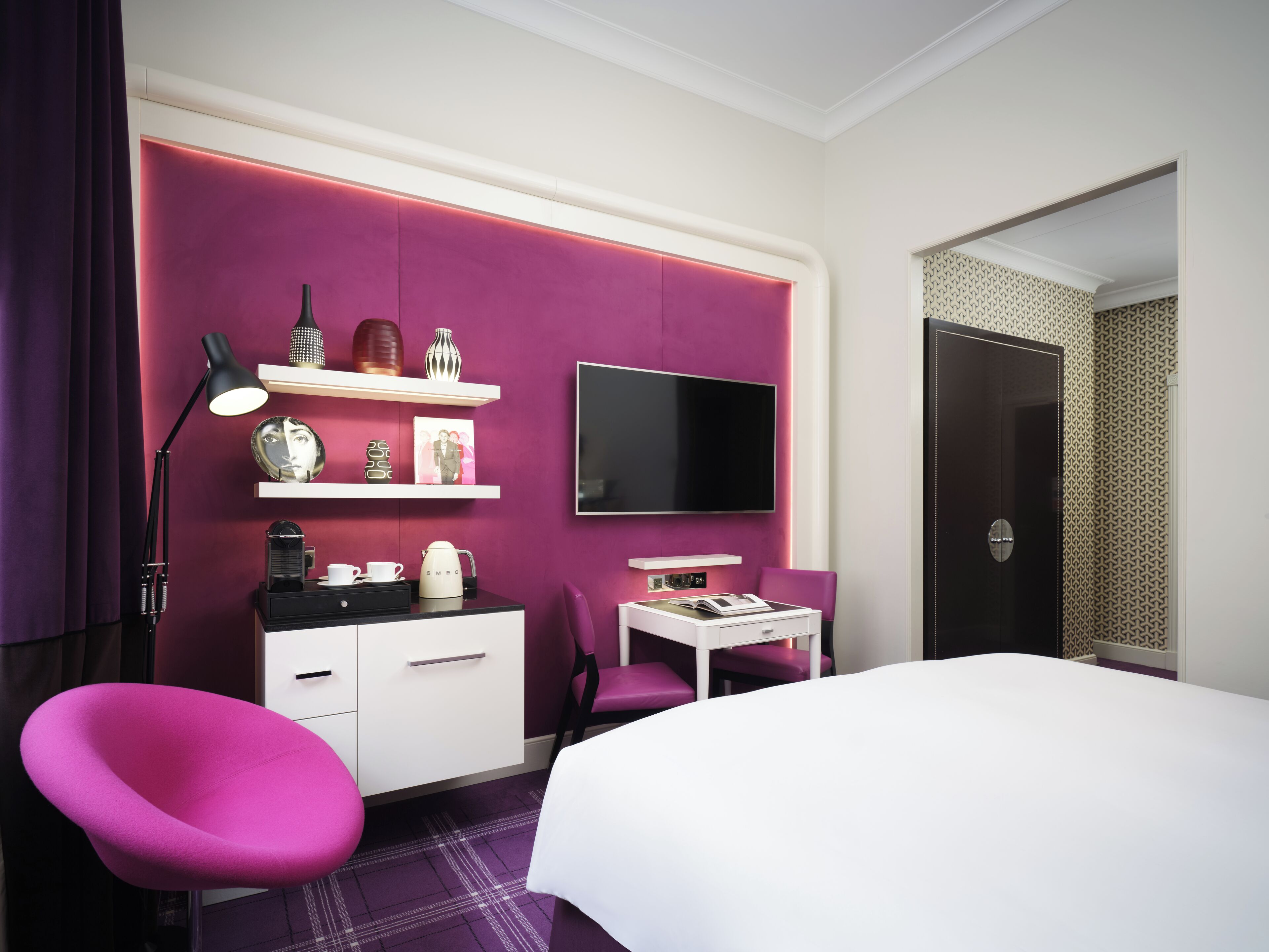 A modern hotel room with a large bed, a desk with a flat-screen TV, a chair, and a vanity with a mirror and decorative items.