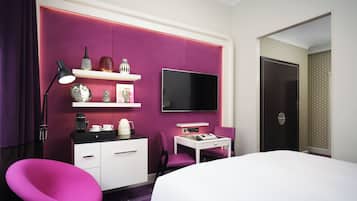 A modern hotel room with a large bed, a desk with a flat-screen TV, a chair, and a vanity with a mirror and decorative items.
