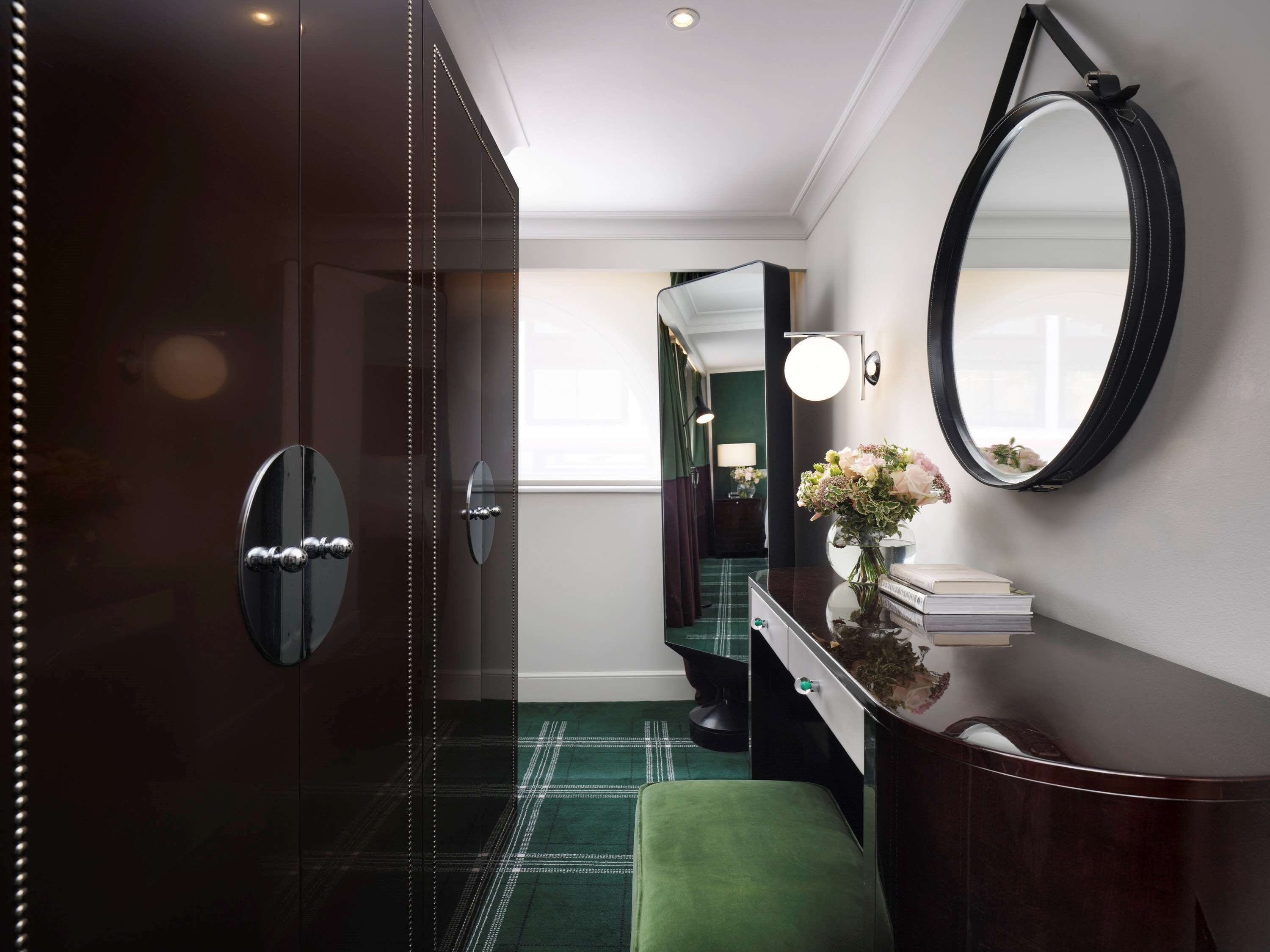 A modern hotel room with a dark wood wardrobe, a green upholstered bench, a vanity with a mirror, and a small table with a vase of flowers.