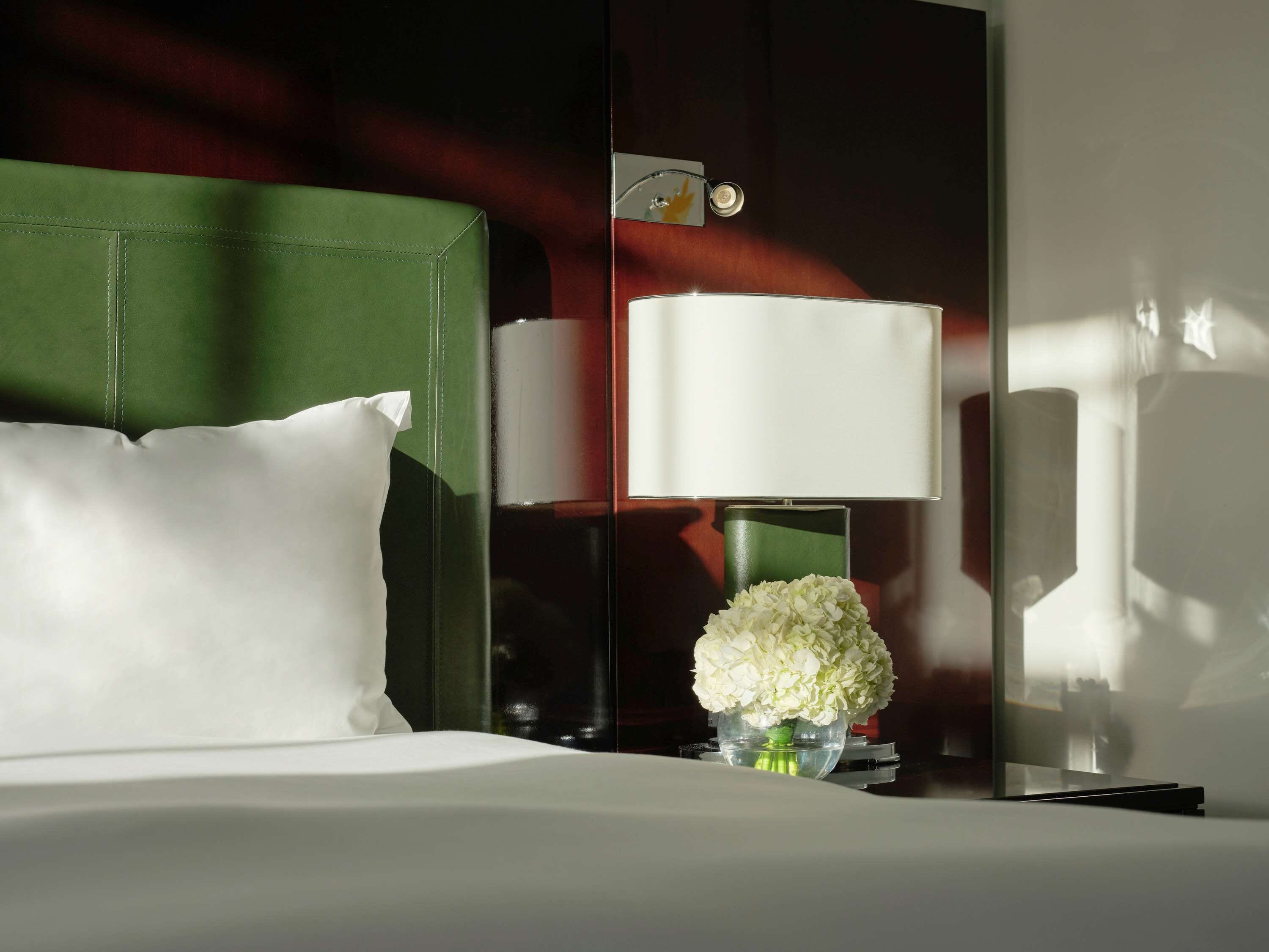 Ein ordentlich bezogenes Bett mit weißen Leinen, einem grünen Kopfteil, einer Nachttischlampe und einer Vase mit weißen Blumen.