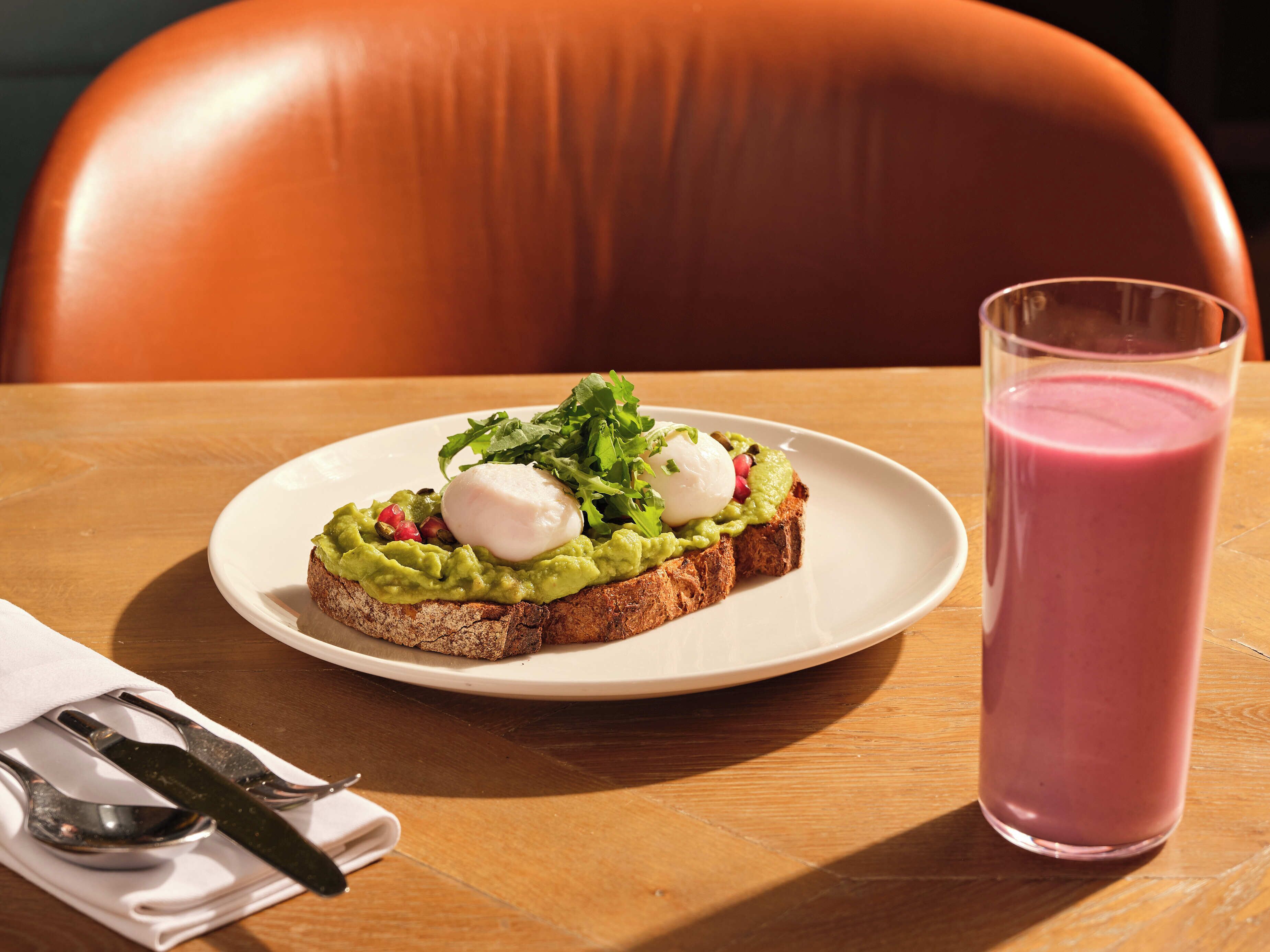 Ein Teller mit einem Sandwich, belegt mit Avocado, Grünzeug und einem pochierten Ei, sowie ein Glas rosa Smoothie auf einem Holztisch.