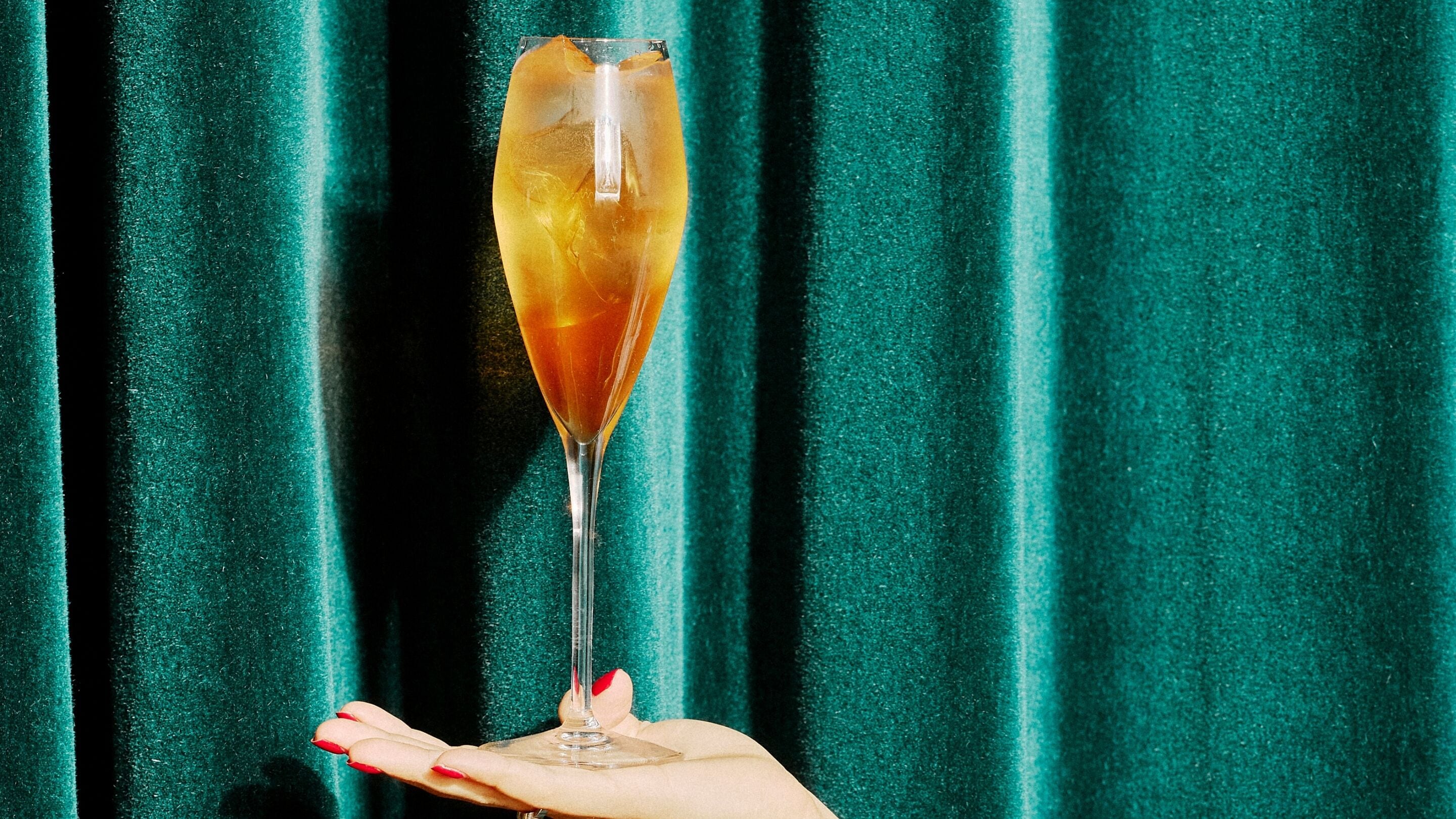 A champagne glass held by a hand against a teal curtain backdrop.
