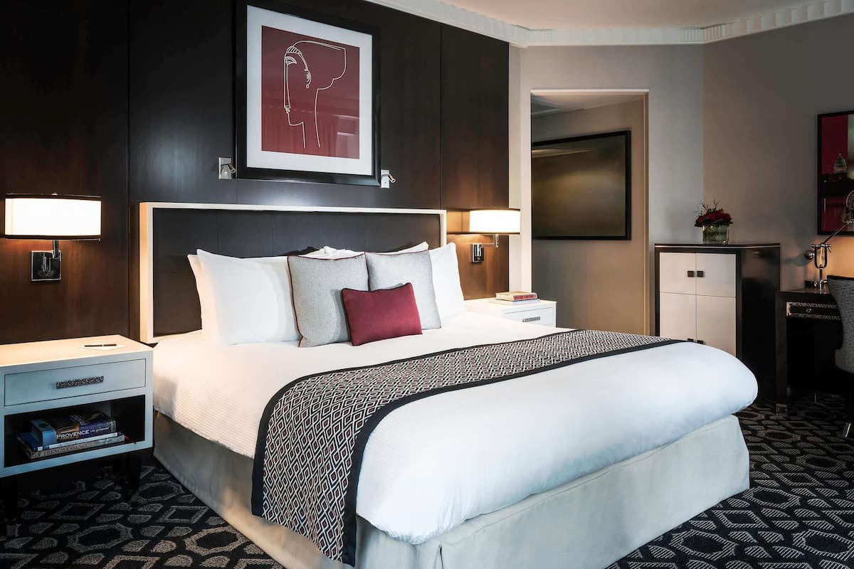 A neatly arranged hotel room with a large bed, two bedside tables, a nightstand, a small cabinet, and a framed artwork above the bed.