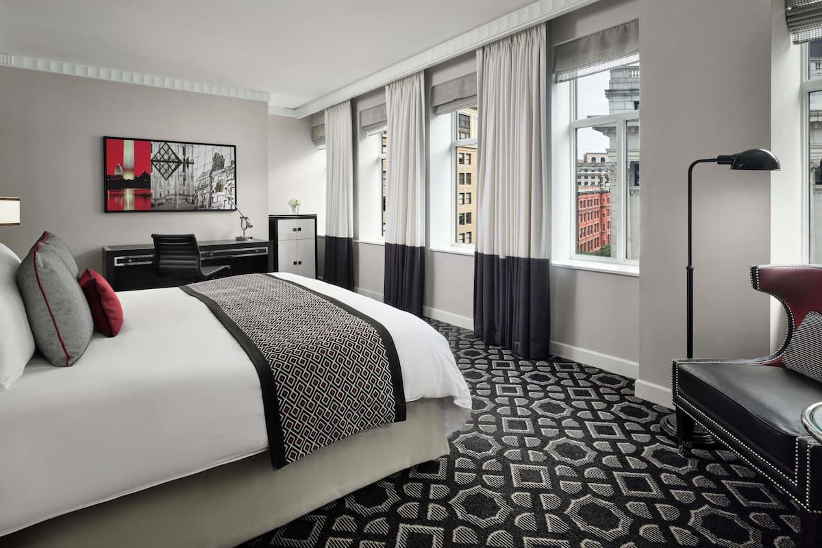 A hotel room with a bed, a desk, a chair, a sofa, a lamp, and a window with curtains.