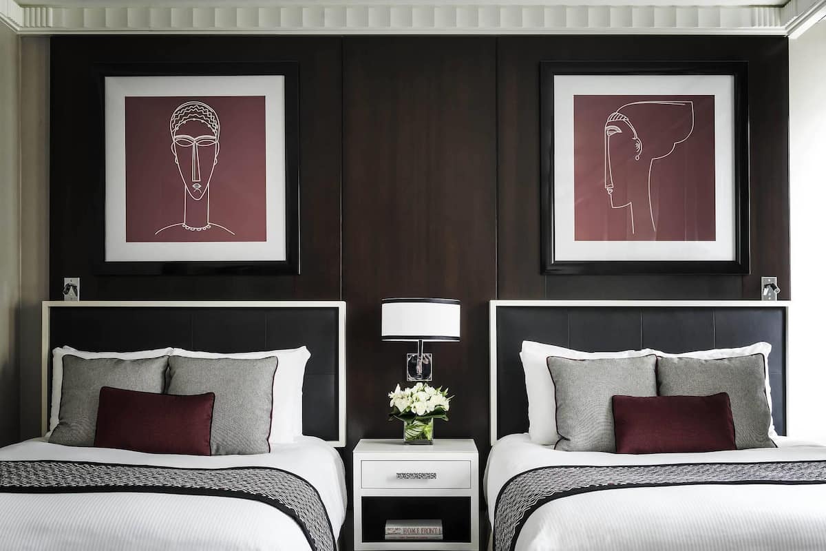 A hotel room with two beds, dark wood paneling, framed artwork above each bed, a bedside table with a lamp and flowers, and a small drawer unit.