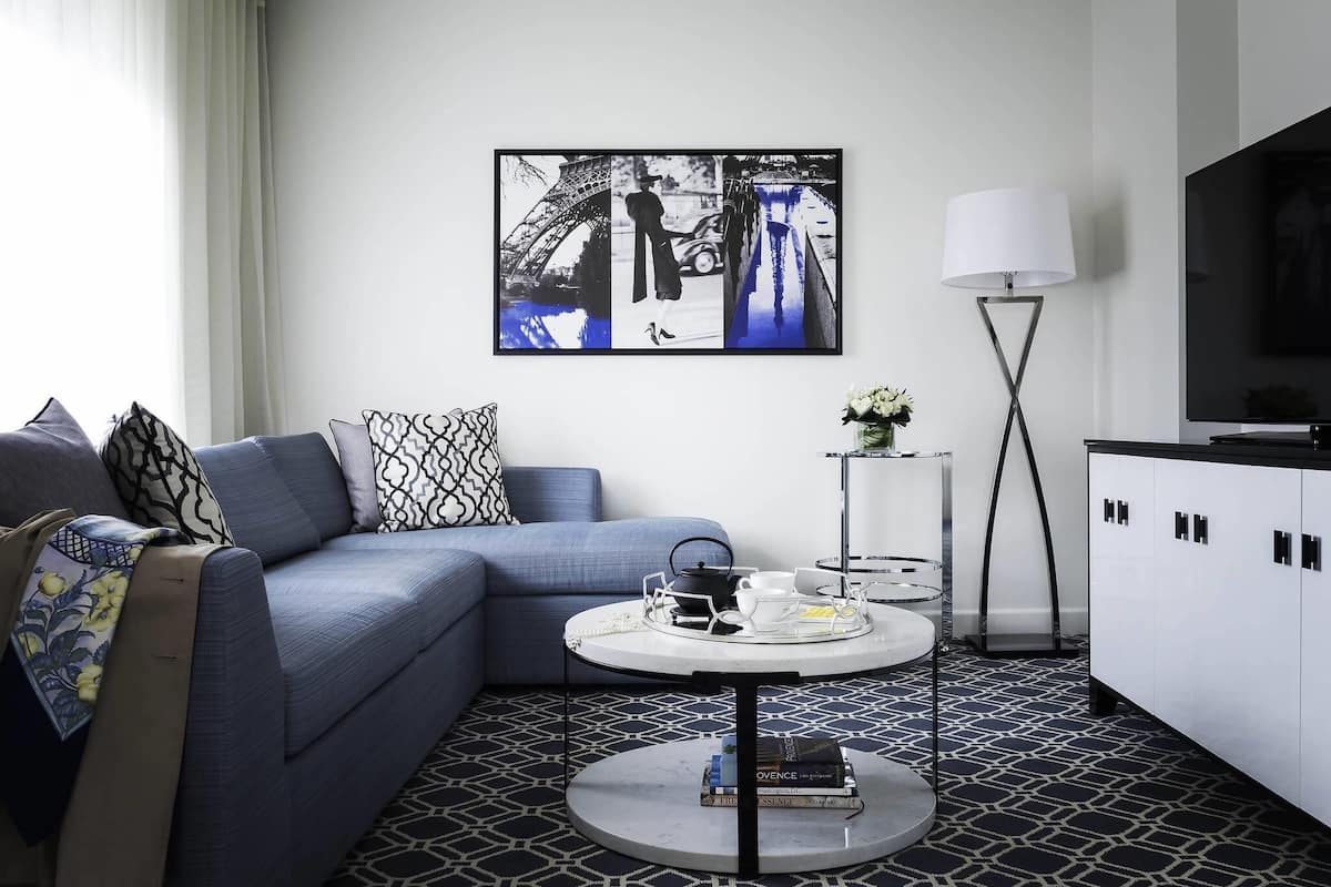 A modern hotel room with a sectional sofa, a round coffee table with a tray, a flat-screen TV, and a large black-and-white artwork of the Eiffel Tower.