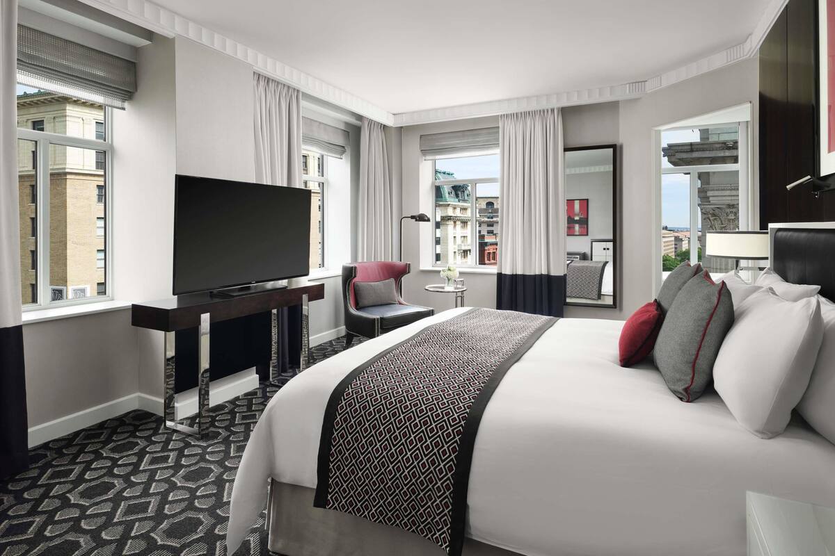 A modern hotel room with a large bed, a flat-screen TV, a desk with a chair, and a view of the city.