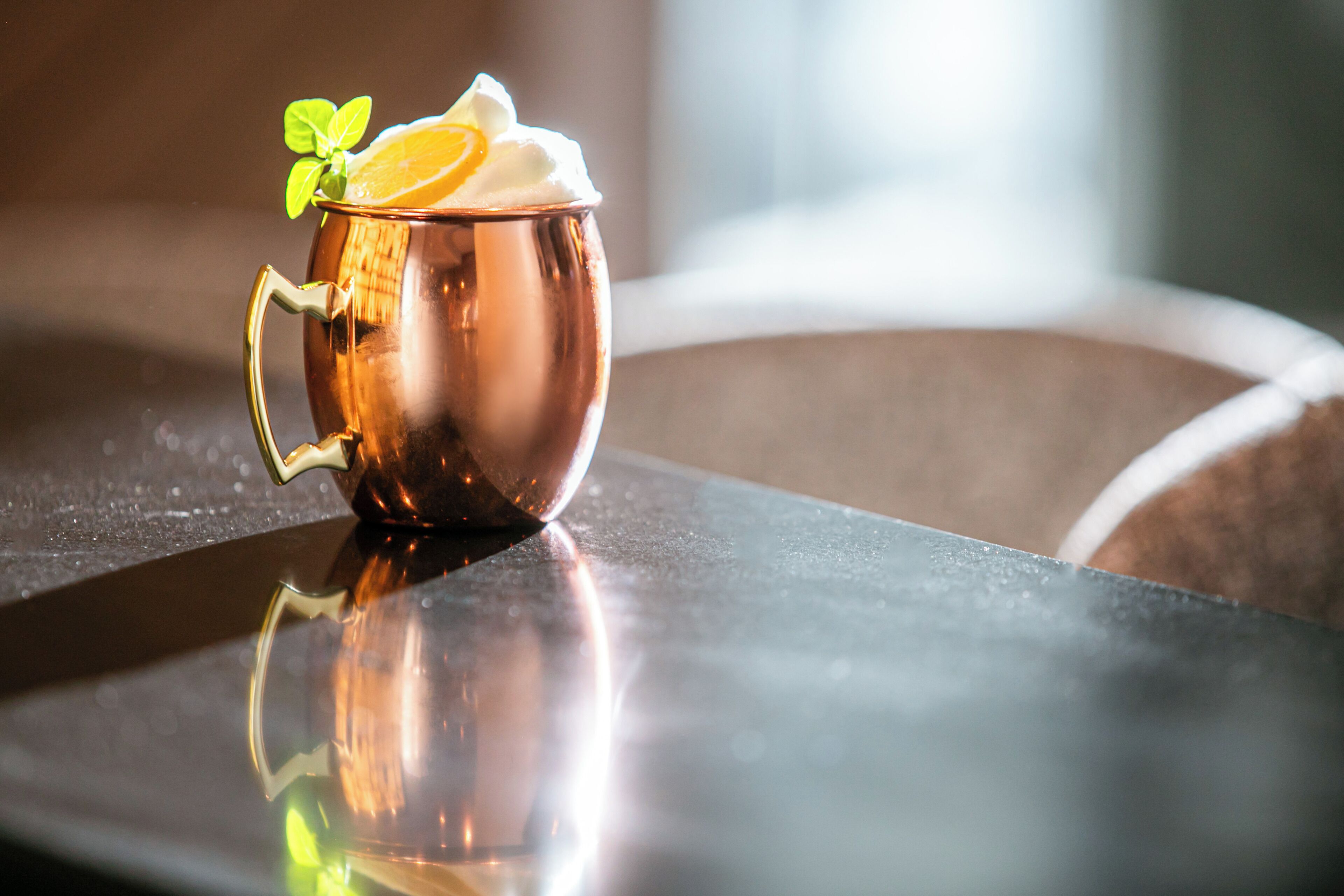 Eine Kupferbecher mit Minzblatt und Orangenscheibe auf einer spiegelnden Oberfläche.