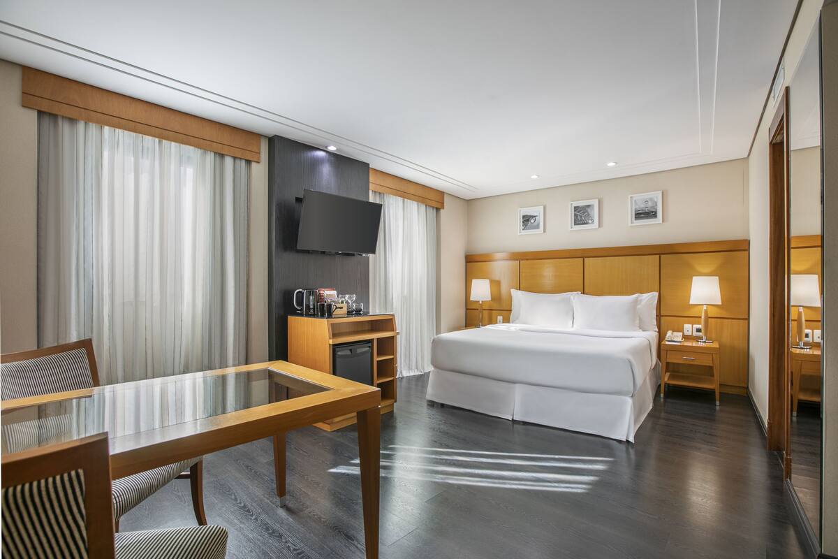 Ein Hotelzimmer mit einem großen Bett, einem Schreibtisch mit Stuhl, einem an der Wand befestigten Fernseher und einem Fenster mit Vorhängen.