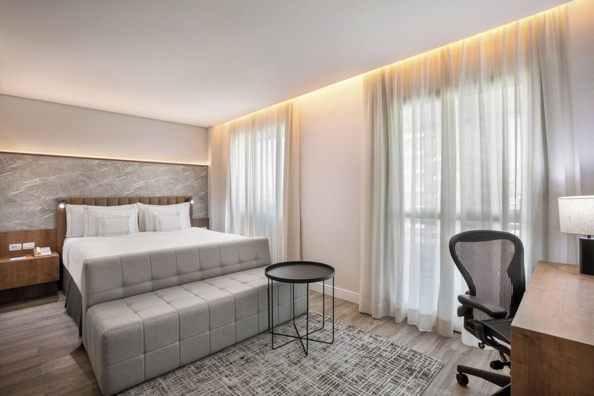Ein modernes Hotelzimmer mit Bett, Schreibtisch, Stuhl, kleinem Tisch und einem großen Fenster mit Vorhängen.