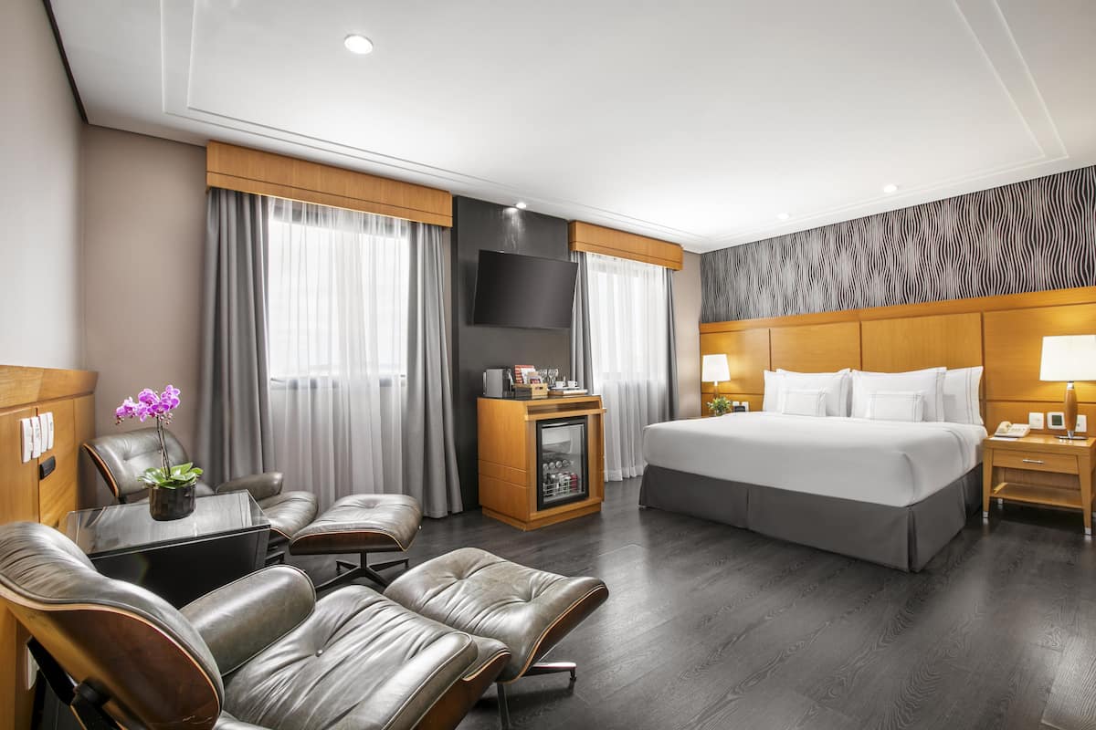 Ein Hotelzimmer mit einem großen Bett, zwei Ledersesseln, einem kleinen Tisch mit einer Pflanze, einer Minibar und einem an der Wand befestigten Fernseher.