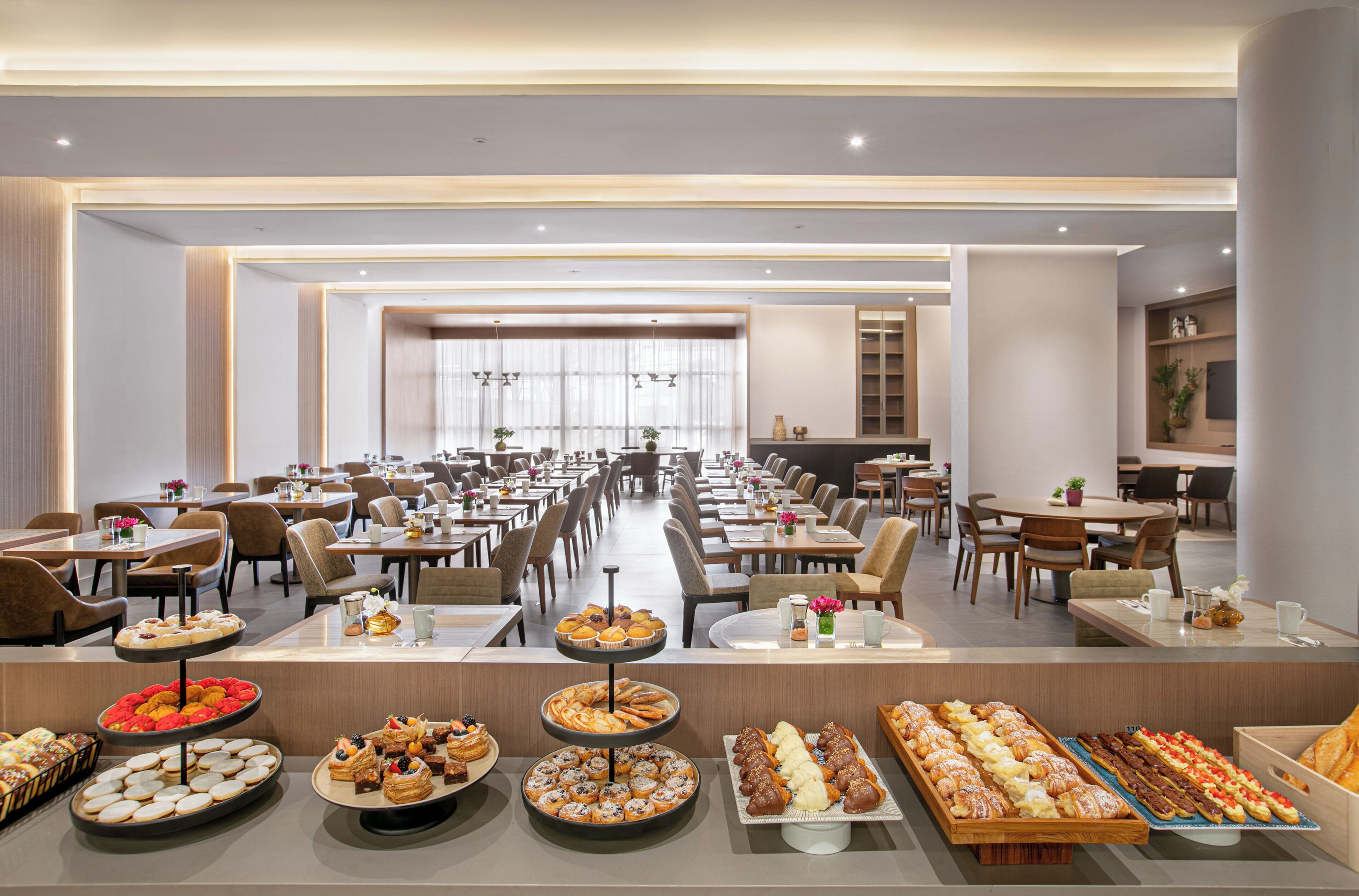 A large dining area with multiple tables and chairs, a buffet with various pastries and desserts, and a long counter with a variety of food items.