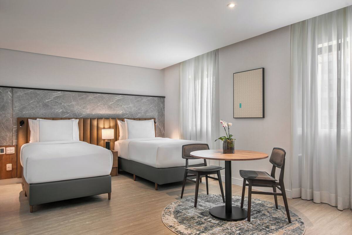 Ein Hotelzimmer mit zwei Betten, einem runden Esstisch, Stühlen, einem kleinen Topfpflanzen und einem gerahmten Bild an der Wand.