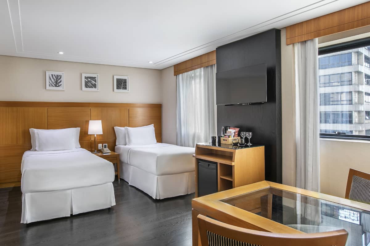 Ein Hotelzimmer mit zwei Betten, einem Schreibtisch, einem Fernseher und einem Fenster mit Blick auf die Stadt.