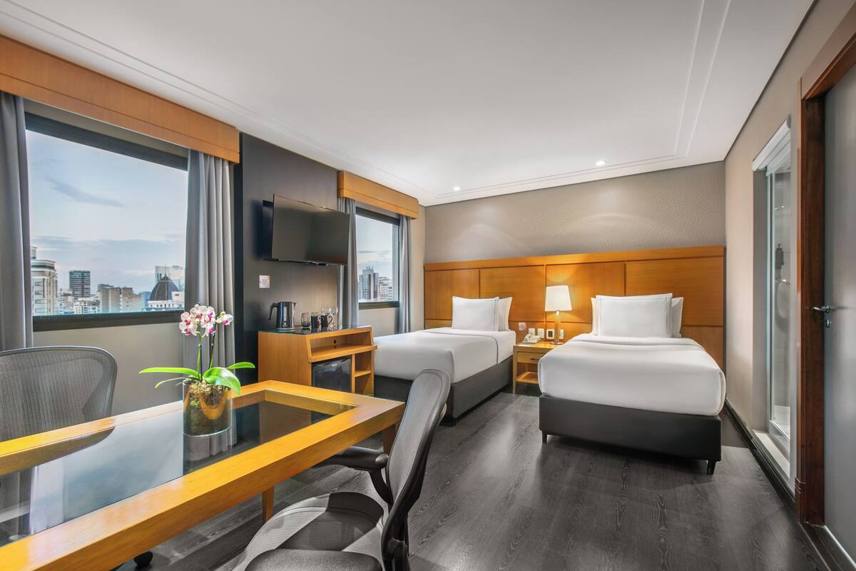 Ein Hotelzimmer mit zwei Betten, einem Schreibtisch, einem Sessel, einem Fernseher und einem Fenster mit Blick auf die Stadt.