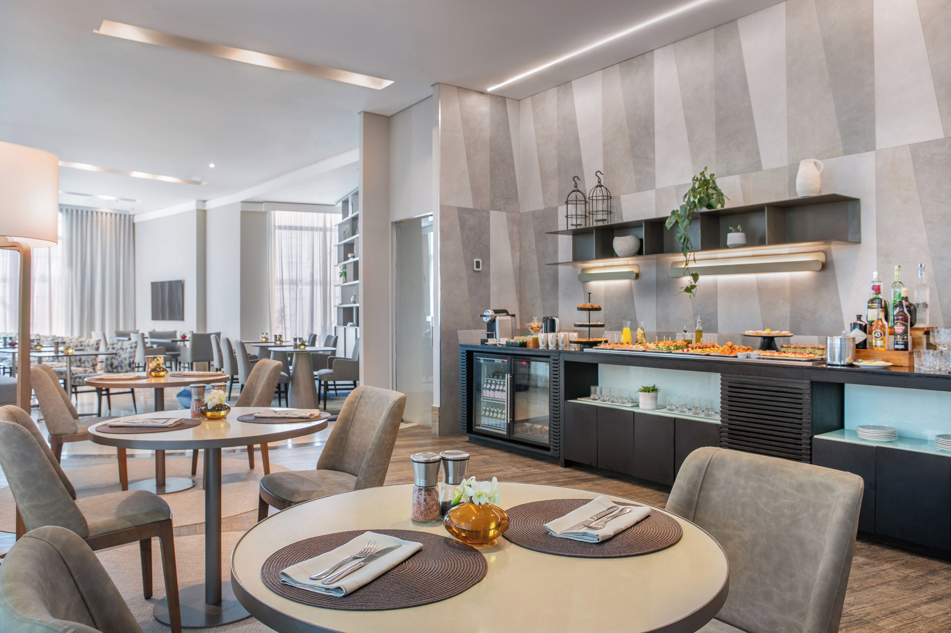 A modern hotel dining area with round tables, comfortable seating, a buffet counter, and a kitchen area.