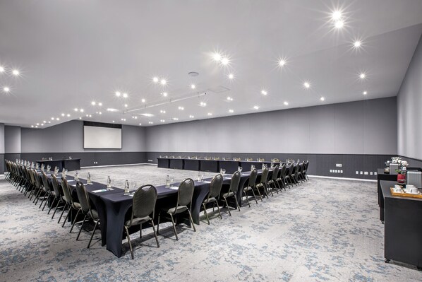 Un amplio salón de conferencias con una larga mesa, sillas y una pantalla de proyector.