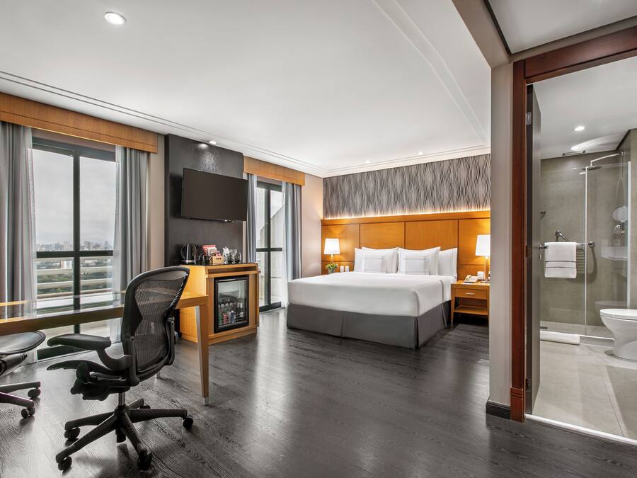 A hotel room with a large bed, a desk with a chair, a TV, and a shower area.