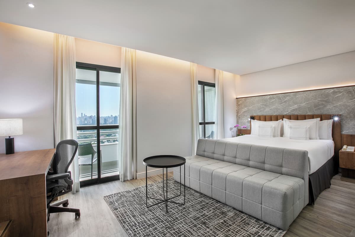 Ein modernes Hotelzimmer mit einem großen Bett, einem Schreibtisch, einem Stuhl und Blick auf die Stadt durch große Fenster.
