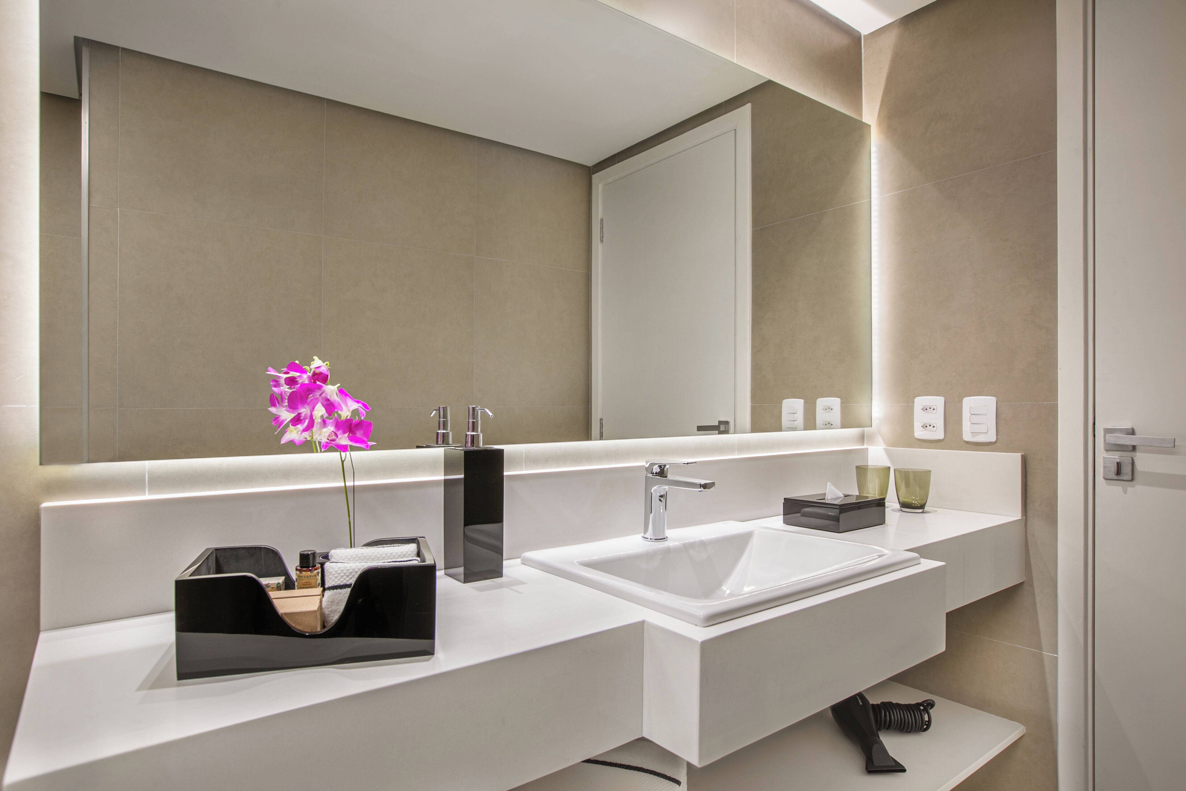 A modern bathroom with a large mirror, white sink, and a black soap dispenser.