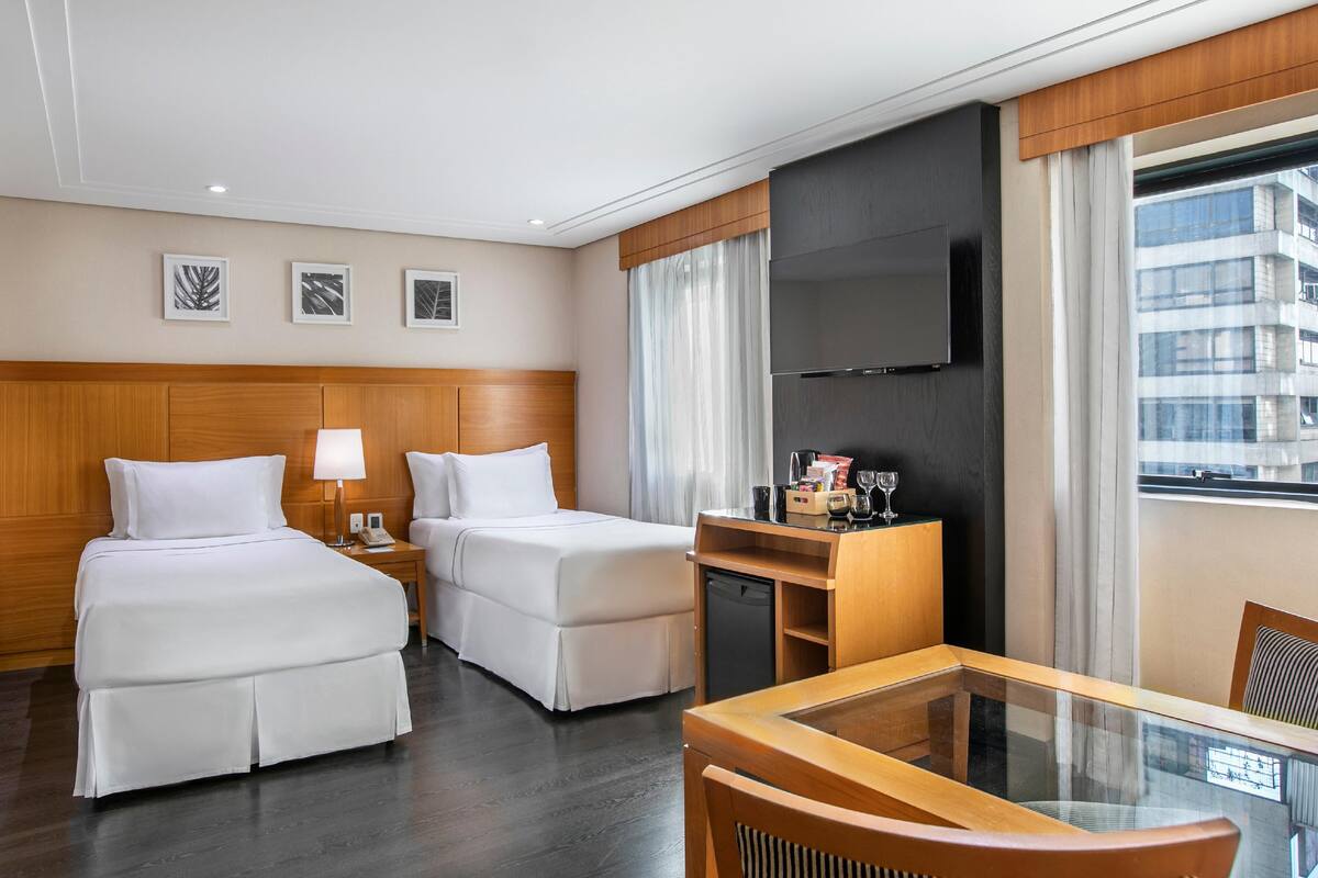 Ein Hotelzimmer mit zwei Betten, einem Schreibtisch, einem Fernseher und Blick auf ein Gebäude.