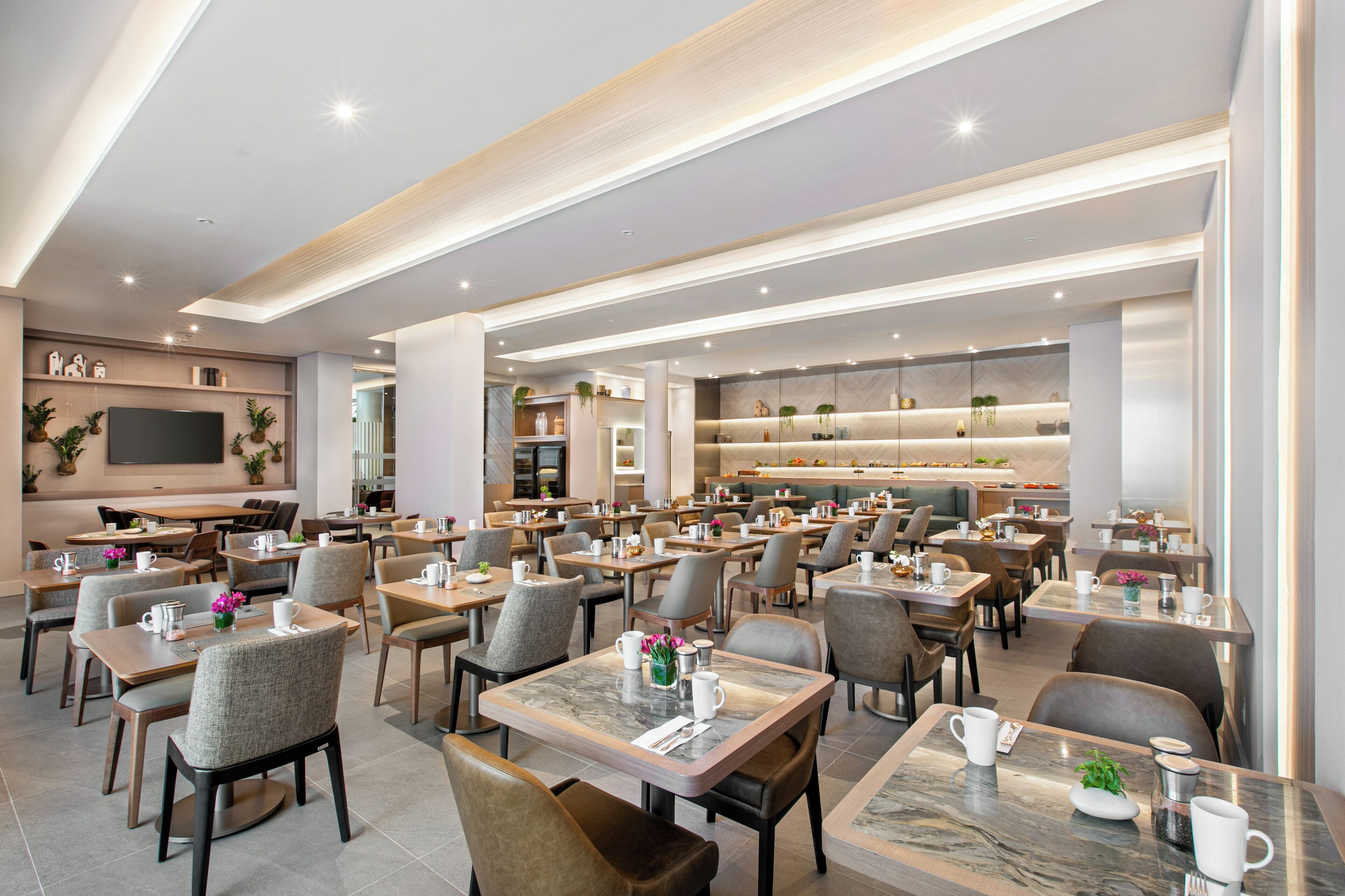 A spacious dining area with multiple tables and chairs, a large flat-screen TV mounted on the wall, and a kitchen area in the background.