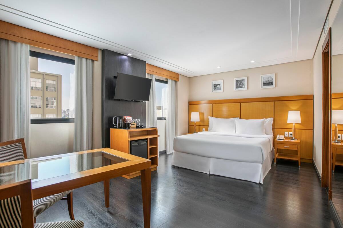 Ein Hotelzimmer mit Bett, Fernseher, Schreibtisch mit Stuhl, Tisch und einem Fenster mit Vorhängen.
