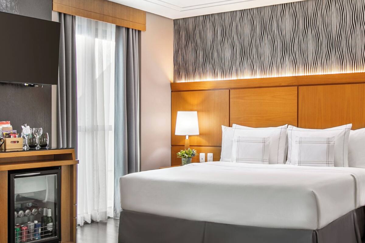 Ein Hotelzimmer mit einem großen Bett, einem Nachttisch mit Lampe, einer Minibar und einem Fernseher.
