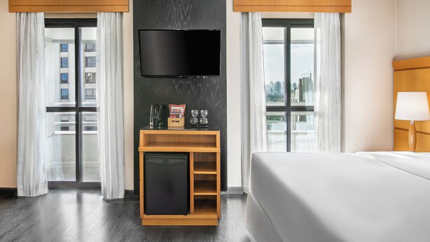 Habitación de hotel con cama, televisor montado en la pared, un pequeño mueble de madera y vistas a la ciudad a través de amplias ventanas.