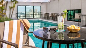 A poolside table with a drink, sunglasses, and a bowl of chips, with a chair and a striped towel nearby.