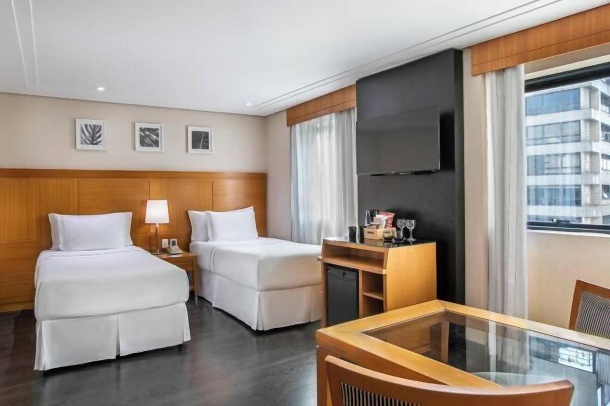 A hotel room with two beds, a desk, a TV, and a window with a city view.
