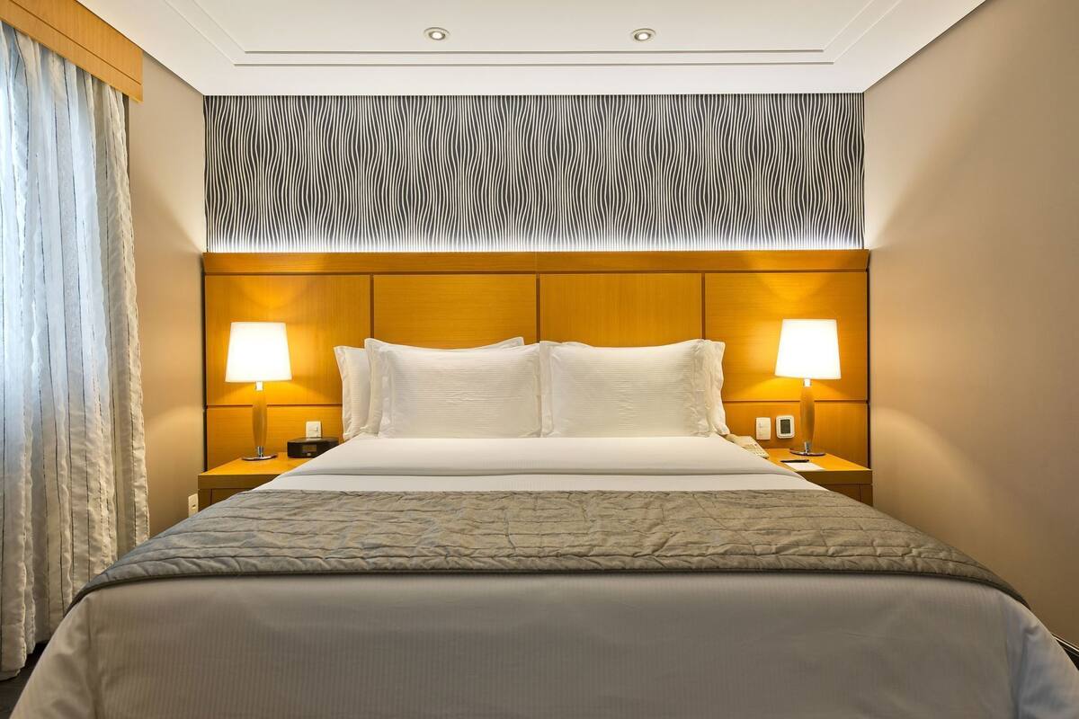 A hotel room with a large bed, two bedside lamps, a patterned headboard, and a curtain on the left.