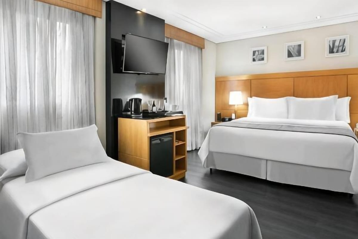 Ein Hotelzimmer mit zwei Betten, einem Fernseher, einem Schreibtisch mit Kaffeemaschine und einem Nachttisch mit Lampe.
