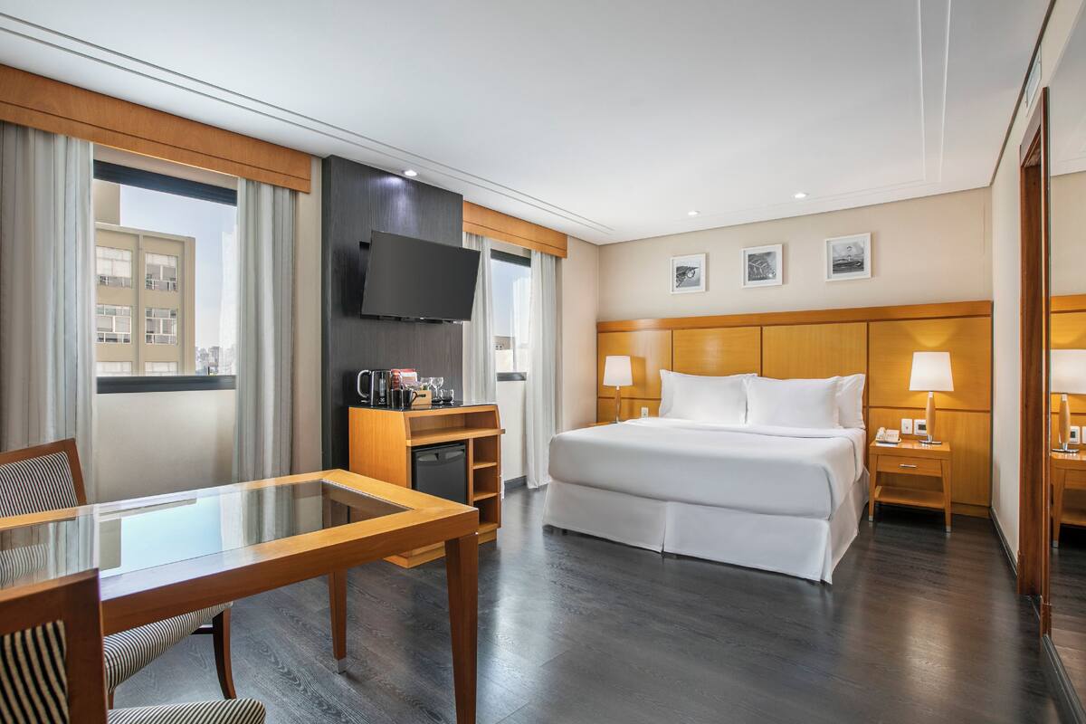 Ein Hotelzimmer mit einem großen Bett, einem an der Wand montierten Fernseher, einem Schreibtisch mit Stuhl und einem Fenster mit Vorhängen.