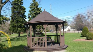 Una pérgola con techo y un banco en su interior, rodeada de césped y árboles.