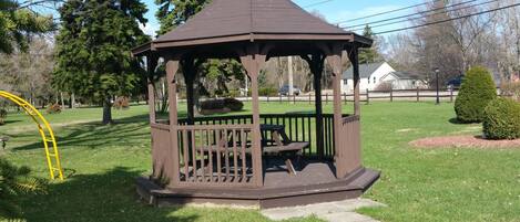 Una pérgola con techo y un banco en su interior, rodeada de césped y árboles.